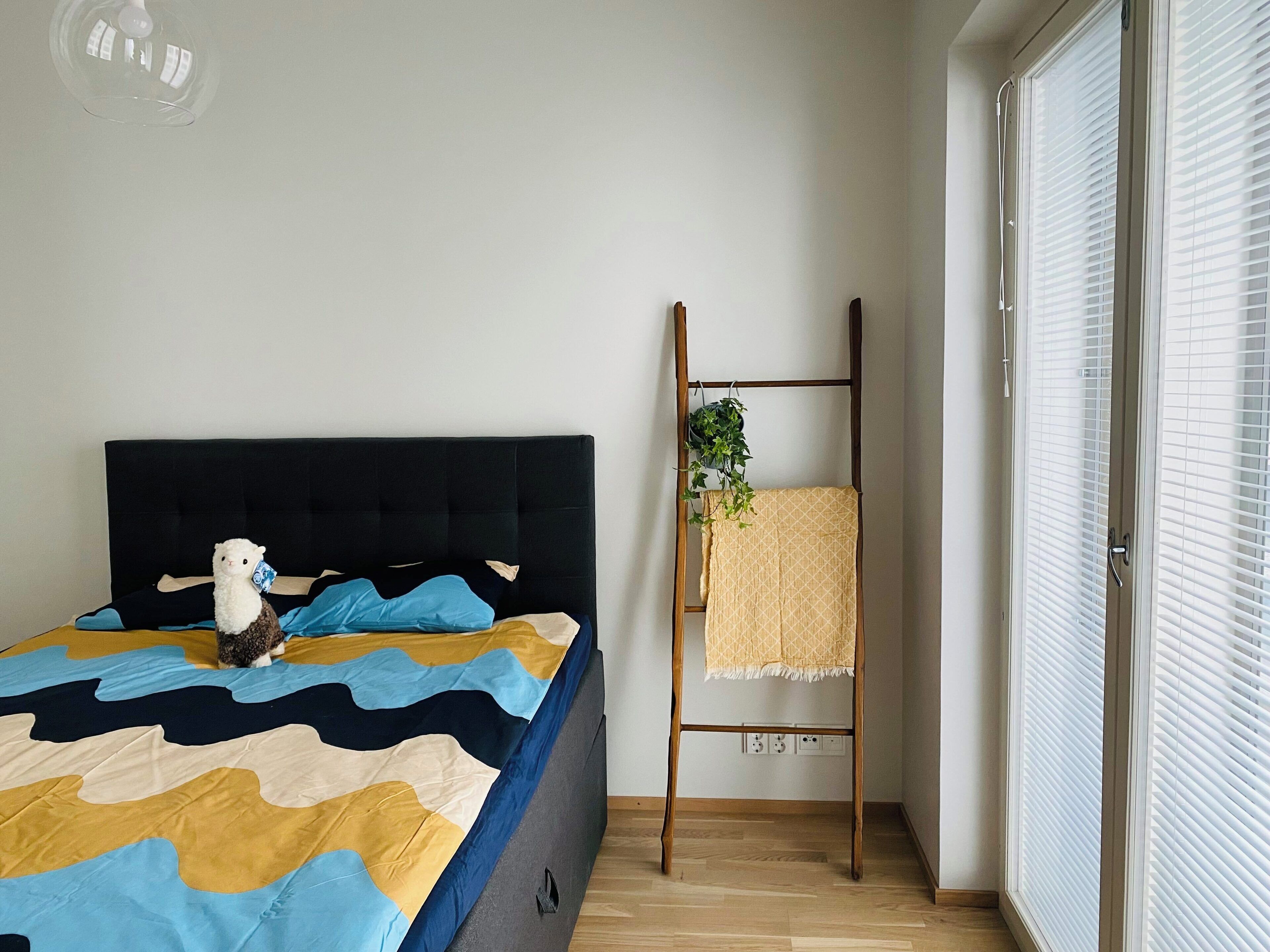 A bedroom with a bed, a small stuffed animal, a wooden ladder with a plant, and a window with blinds.