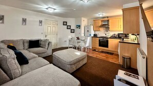 Un espacio habitable compacto con un sofá gris, una mesa de comedor con sillas, una kitchenette con armarios de madera y una puerta blanca.