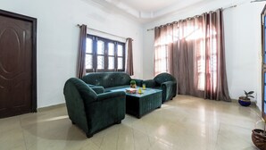 A spacious living room with a dark green sofa set, a coffee table, and a large window with curtains. - Serene 2-bedroom with AC and Kitchen in vibrant Dehradun (Dehradun)