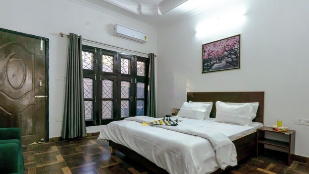 A bedroom with a large bed, a wooden floor, a green chair, a nightstand, and a window with curtains. - Serene 2-bedroom with AC and Kitchen in vibrant Dehradun (Dehradun)