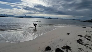 Ranta, jossa henkilö kahlaa matalassa vedessä, kauempana näkyvät vuoret ja pilvinen taivas.