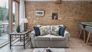 A cozy living area with a stone wall, a sofa with cushions, a small table with a lamp, and a view through a glass door. -  Beautiful one bedroom barn in the rural hamlet of North Wraxall (North Wraxall)