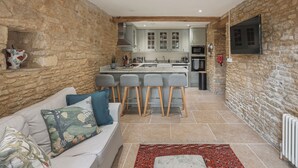 A stone-walled room with a sofa, a red patterned rug, and a kitchen area with a bar stool set. -  Beautiful one bedroom barn in the rural hamlet of North Wraxall (North Wraxall)
