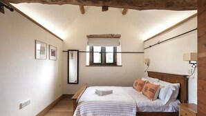 A bedroom with a wooden bed, a window with a white blind, a bedside table, and a wall-mounted lamp. -  Beautiful one bedroom barn in the rural hamlet of North Wraxall (North Wraxall)