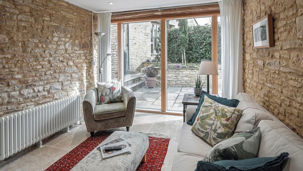 A living room with a stone wall, a sofa, a chair, a red rug, and a view through a glass door to an outdoor area. -  Beautiful one bedroom barn in the rural hamlet of North Wraxall (North Wraxall)