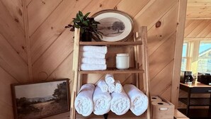 Une étagère en bois avec des serviettes, un tableau encadré et une petite plante en pot, dans une pièce aux murs en bois et dotée d’une fenêtre.