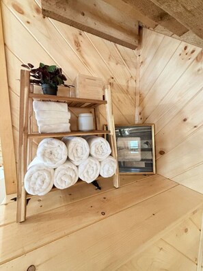 A wooden shelf with towels, a potted plant, and a framed picture in a room with wooden walls and sloped ceilings. - The Heirloom Barn - – Sleep Inside Living History (Bel Air)