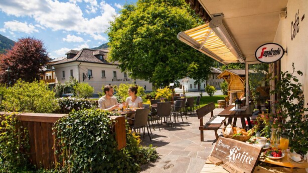 An outdoor café with tables and chairs, surrounded by greenery and a building in the background. - Ferienhof Alte Post (Gitschtal)
