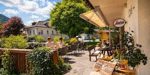 An outdoor café with tables and chairs, surrounded by greenery and a building in the background. - Ferienhof Alte Post (Gitschtal)