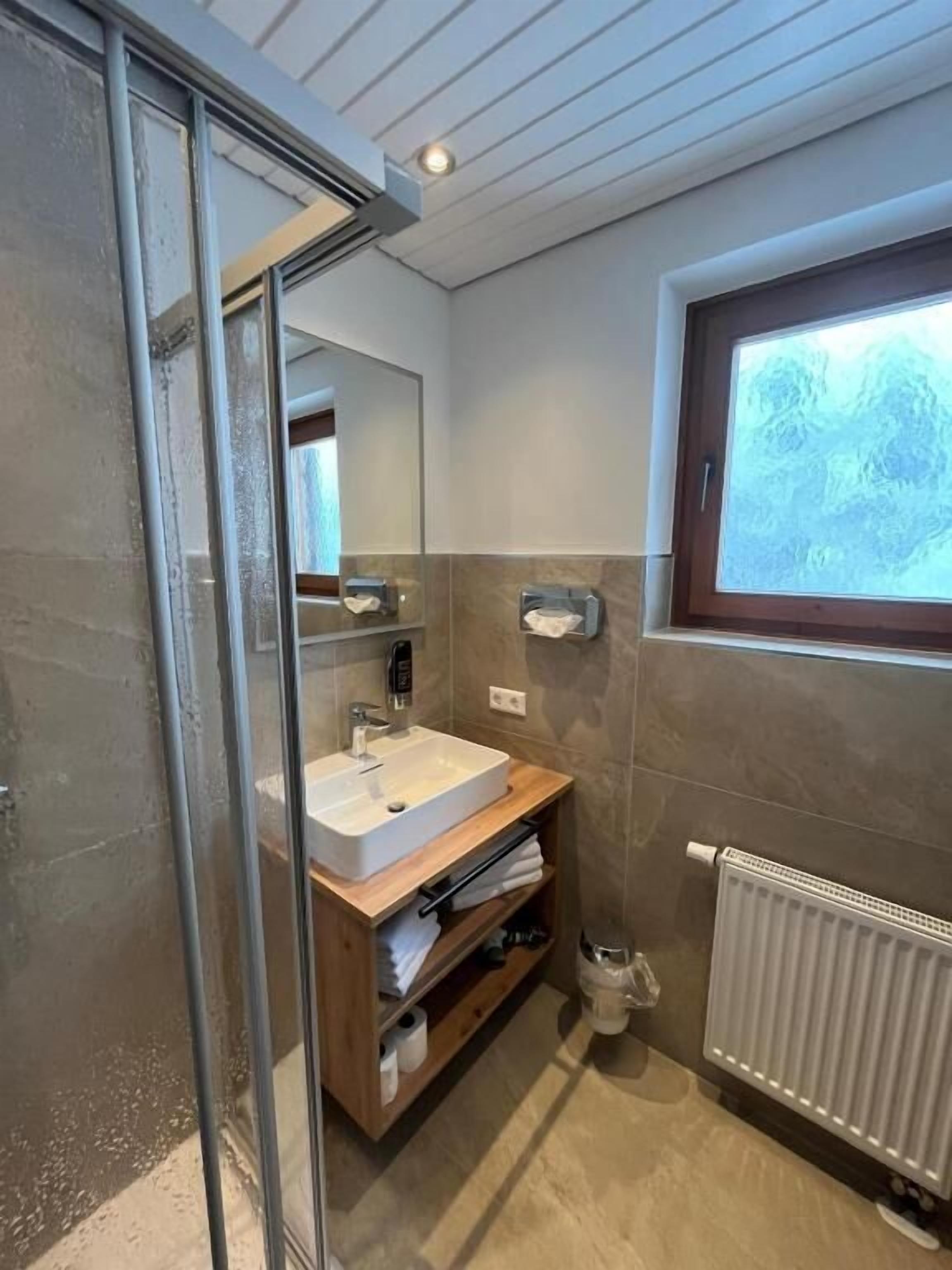 Ein modernes Badezimmer mit Duschkabine aus Glas, einem Holzlagerflächen, einem weißen Waschbecken und einem Fenster mit Blick auf Bäume.