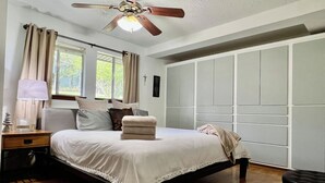 A bedroom with a bed, a nightstand, a lamp, a ceiling fan, and a view of the outdoors through a window.