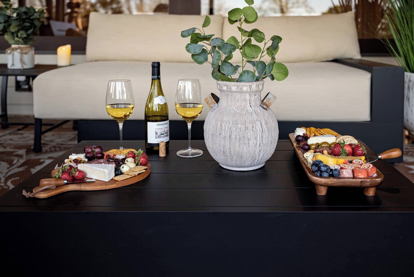 A dining table with a wine bottle, glasses, a vase with green leaves, and a fruit and cheese board.
