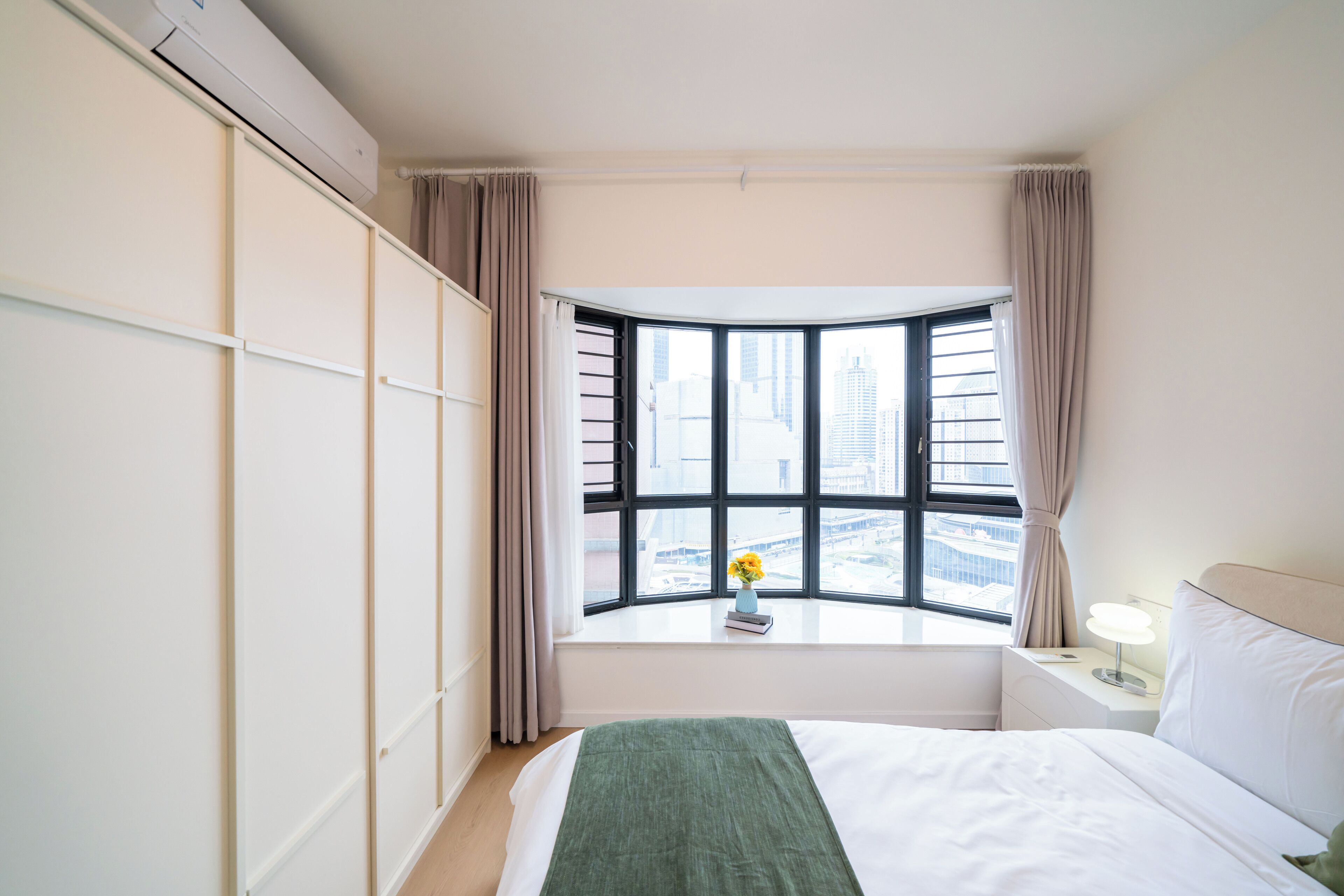 A hotel room with a bed, a nightstand, a window with a view of buildings, and a wall-mounted air conditioner.