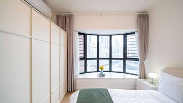 A hotel room with a bed, a nightstand, a window with a view of buildings, and a wall-mounted air conditioner.