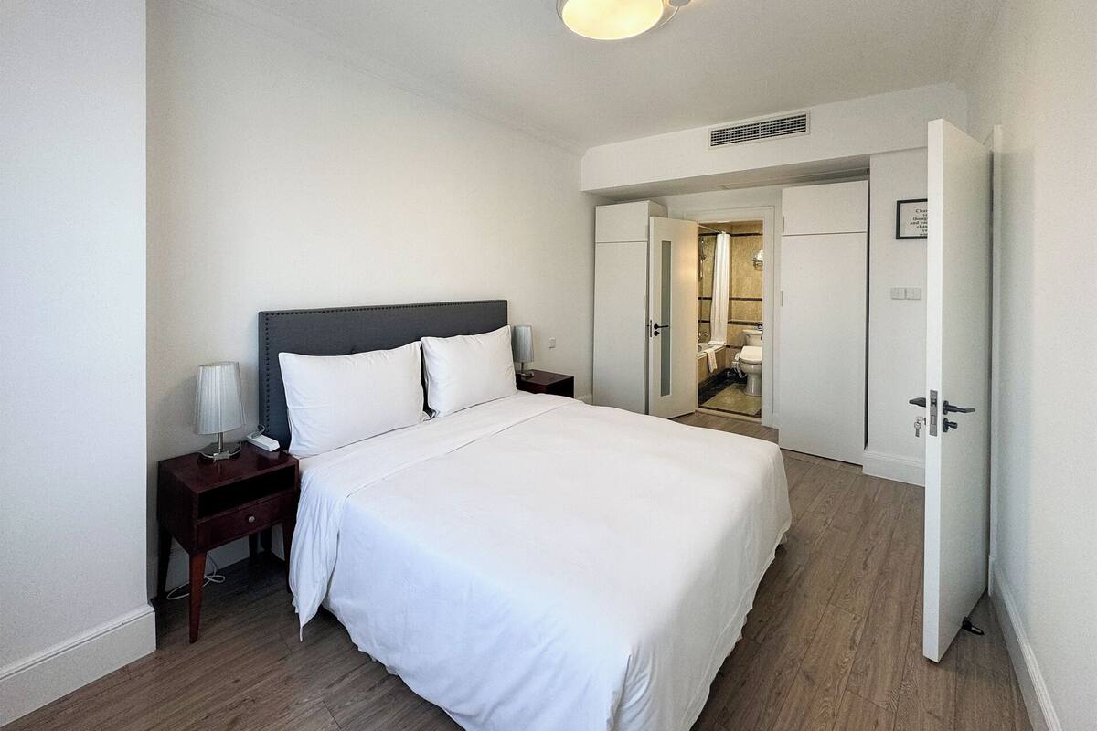 Una habitación de hotel con cama, mesita de noche, lámpara y una puerta abierta que da a un baño.