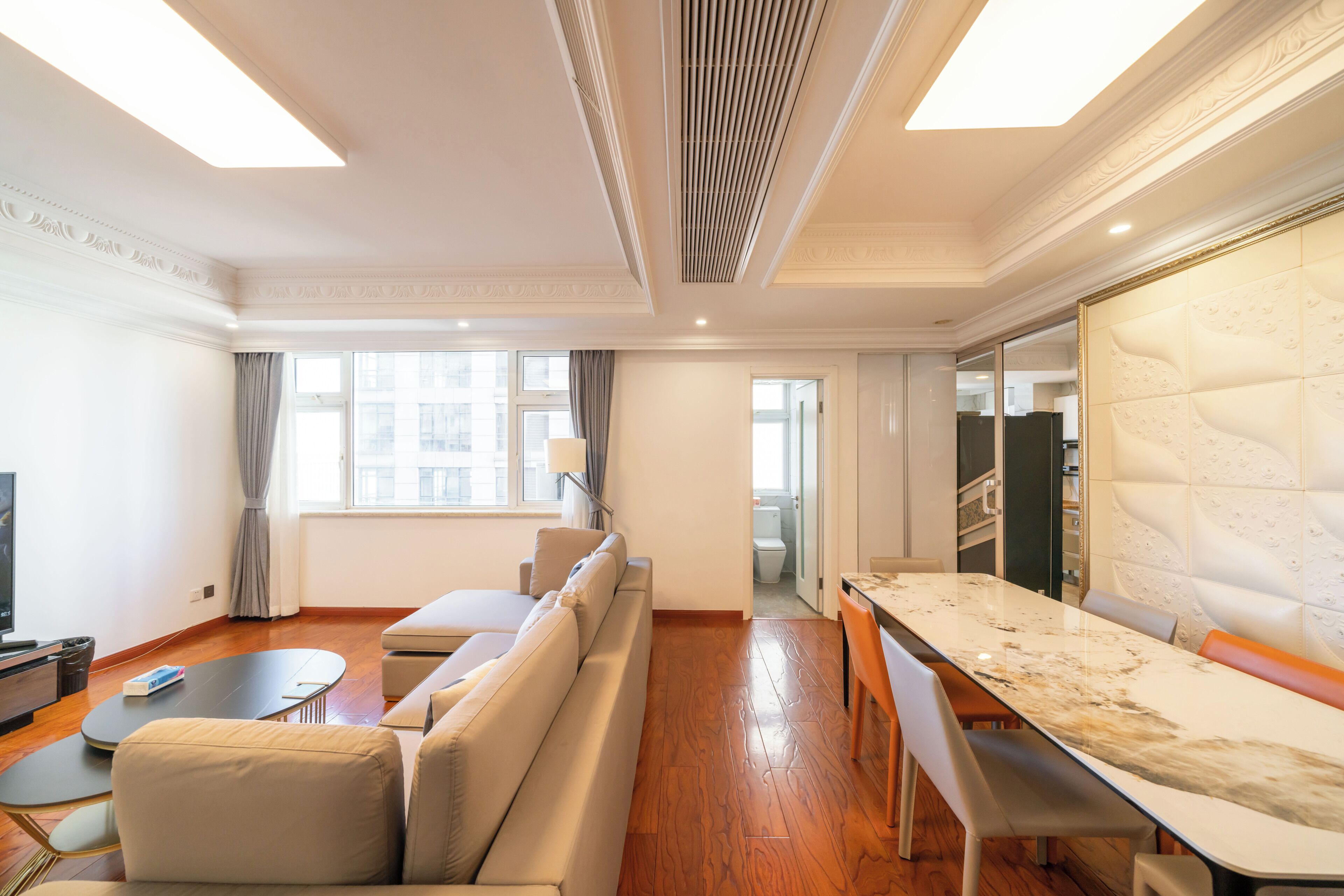 A modern hotel room with a sofa, a dining table, a TV, and a large window.
