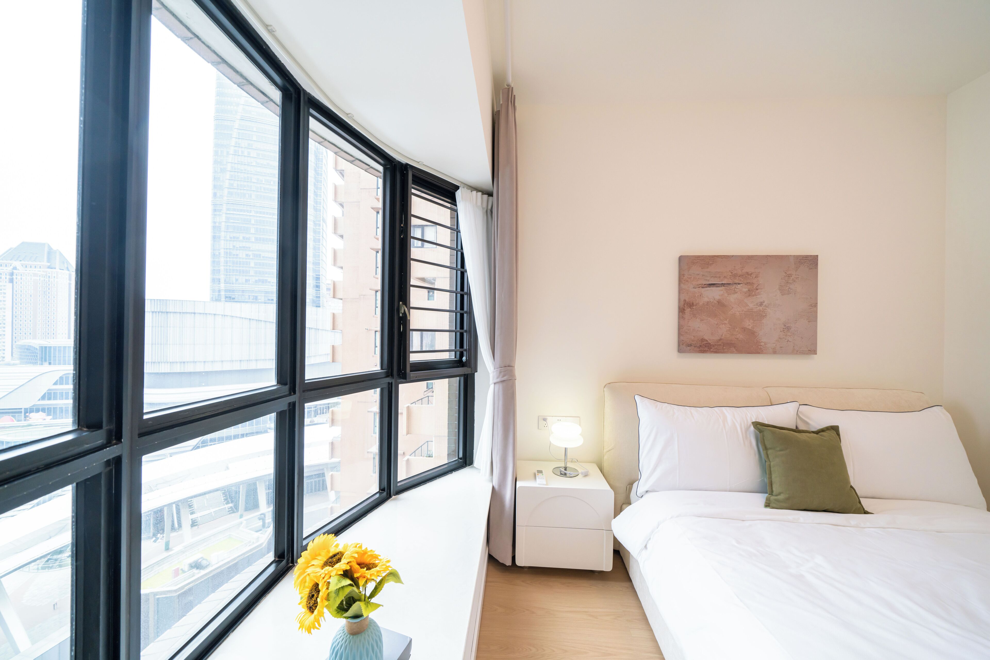 Une chambre d’hôtel avec une grande fenêtre, un lit, une table de chevet, une lampe et un tableau au mur.