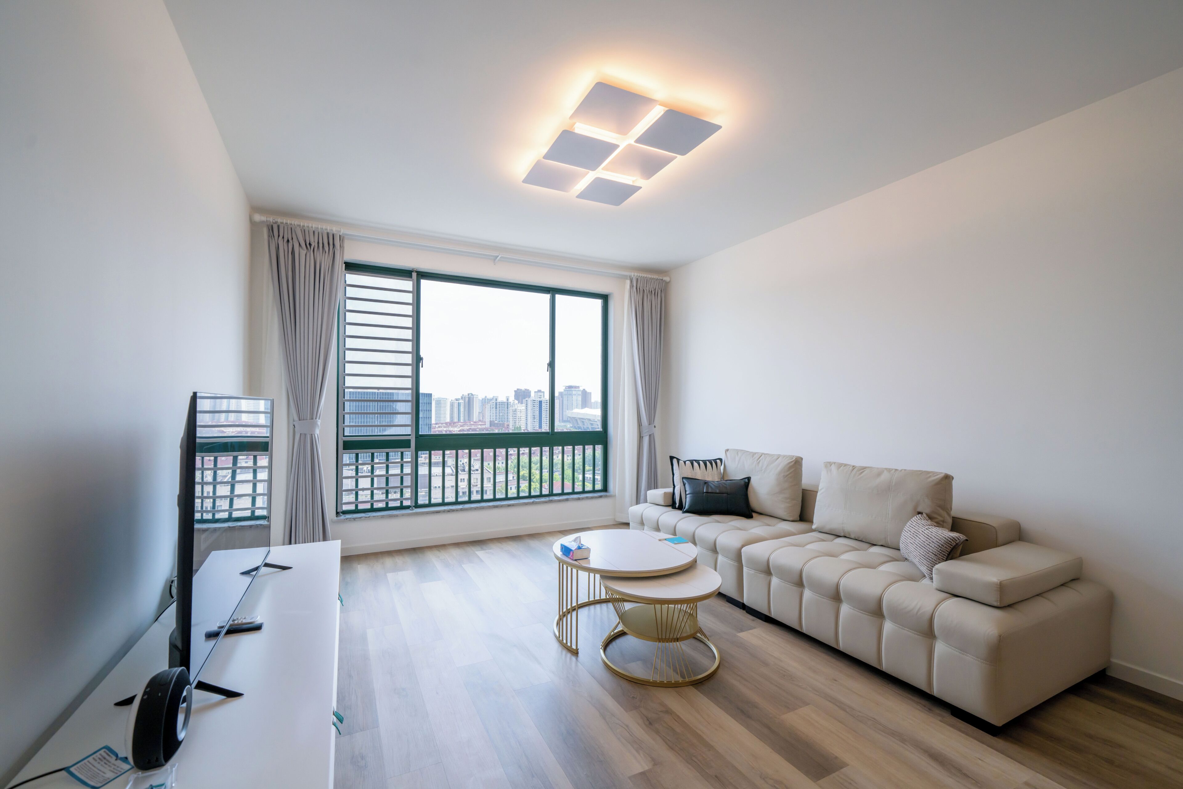 A modern living room with a sofa, a small round table, a flat-screen TV, and a large window with a city view.