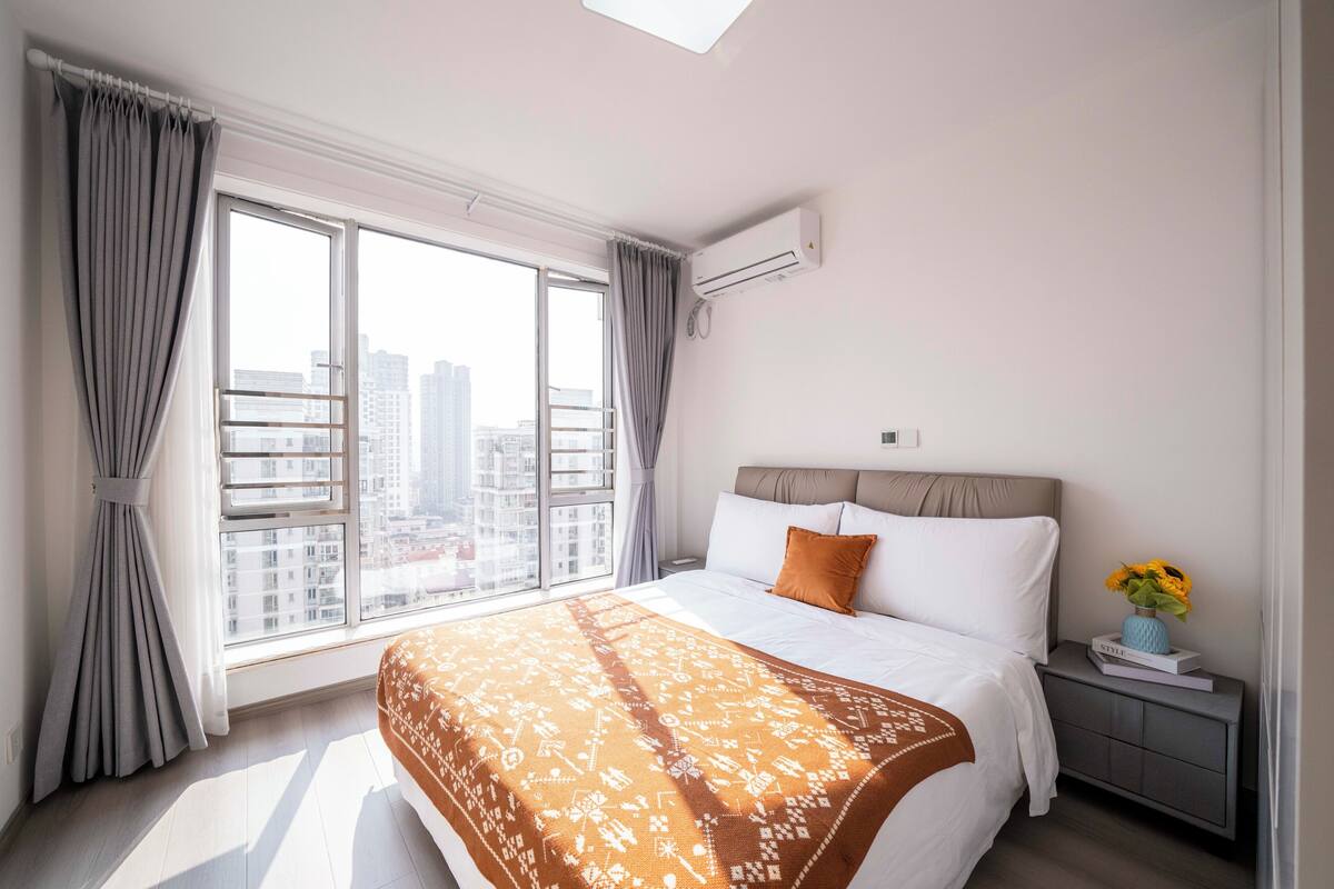 Un dormitorio con un ventanal grande, una cama, una mesita de noche con un florero y un equipo de aire acondicionado.