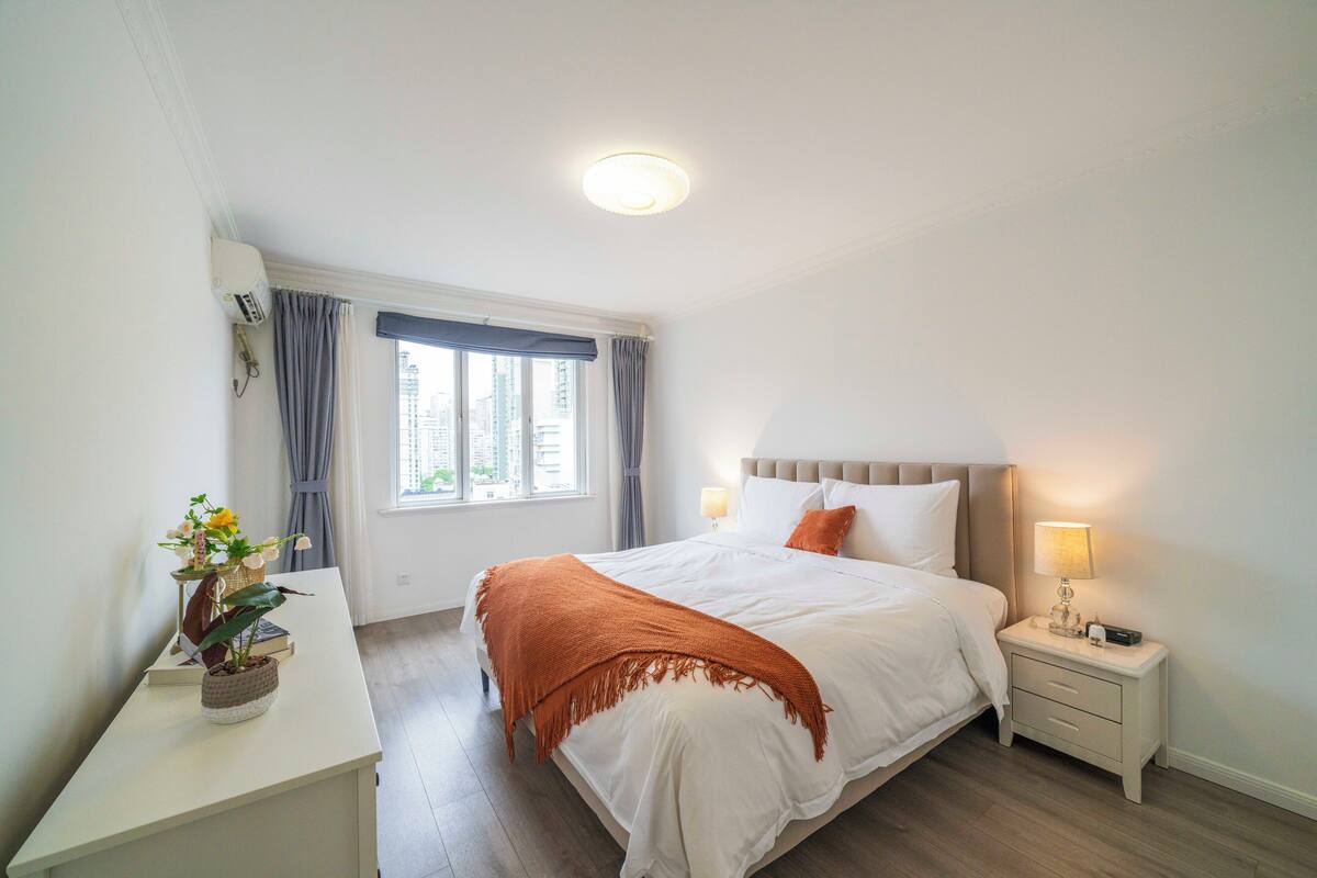 Un dormitorio con cama, mesitas de noche, una ventana con cortinas y una planta en un escritorio.