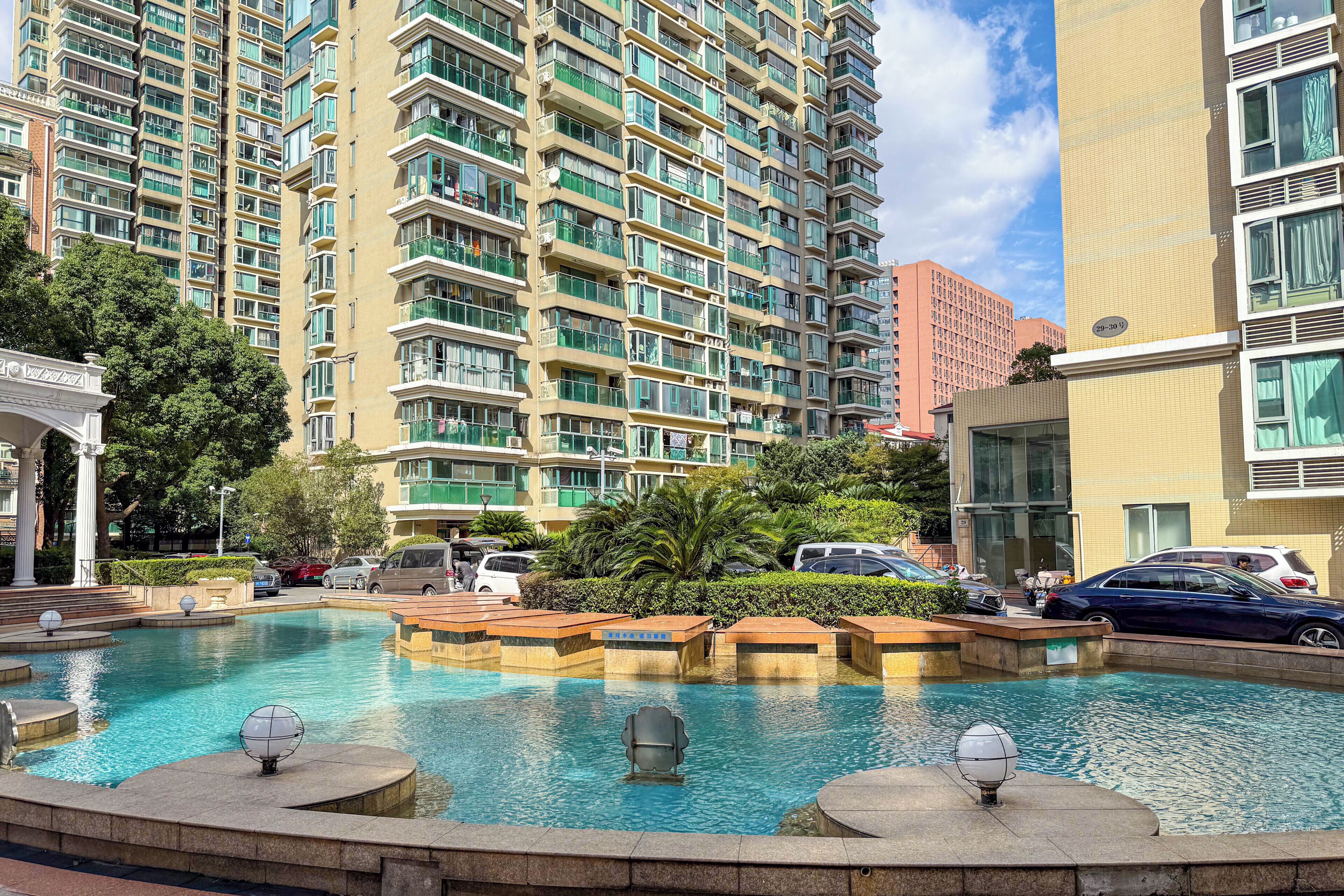 Una piscina con un sillón sumergido, rodeada de vegetación y edificios modernos.