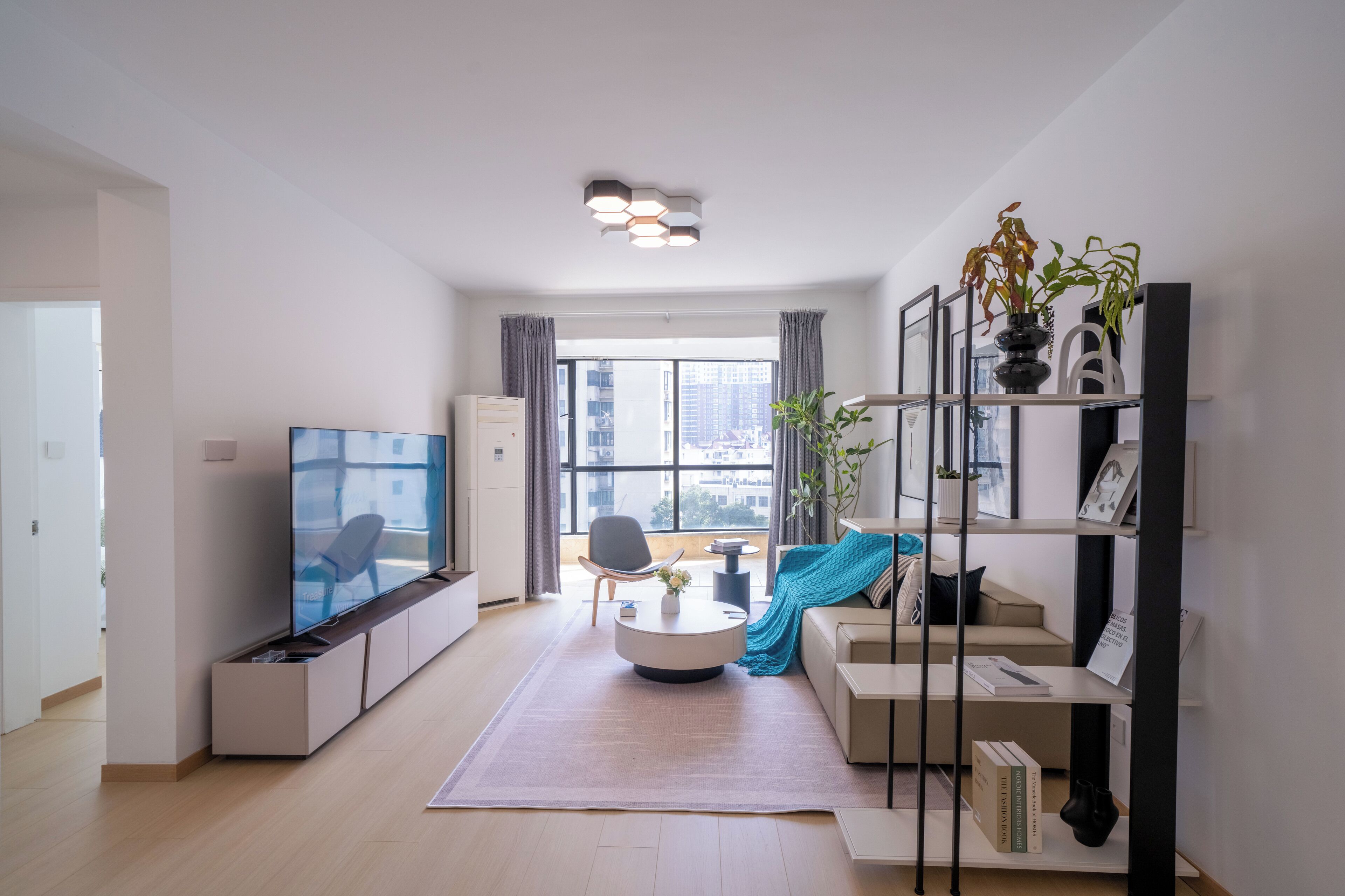 A modern living room with a flat-screen TV, a round coffee table, a sofa, a bookshelf, and a large window with a view of buildings.