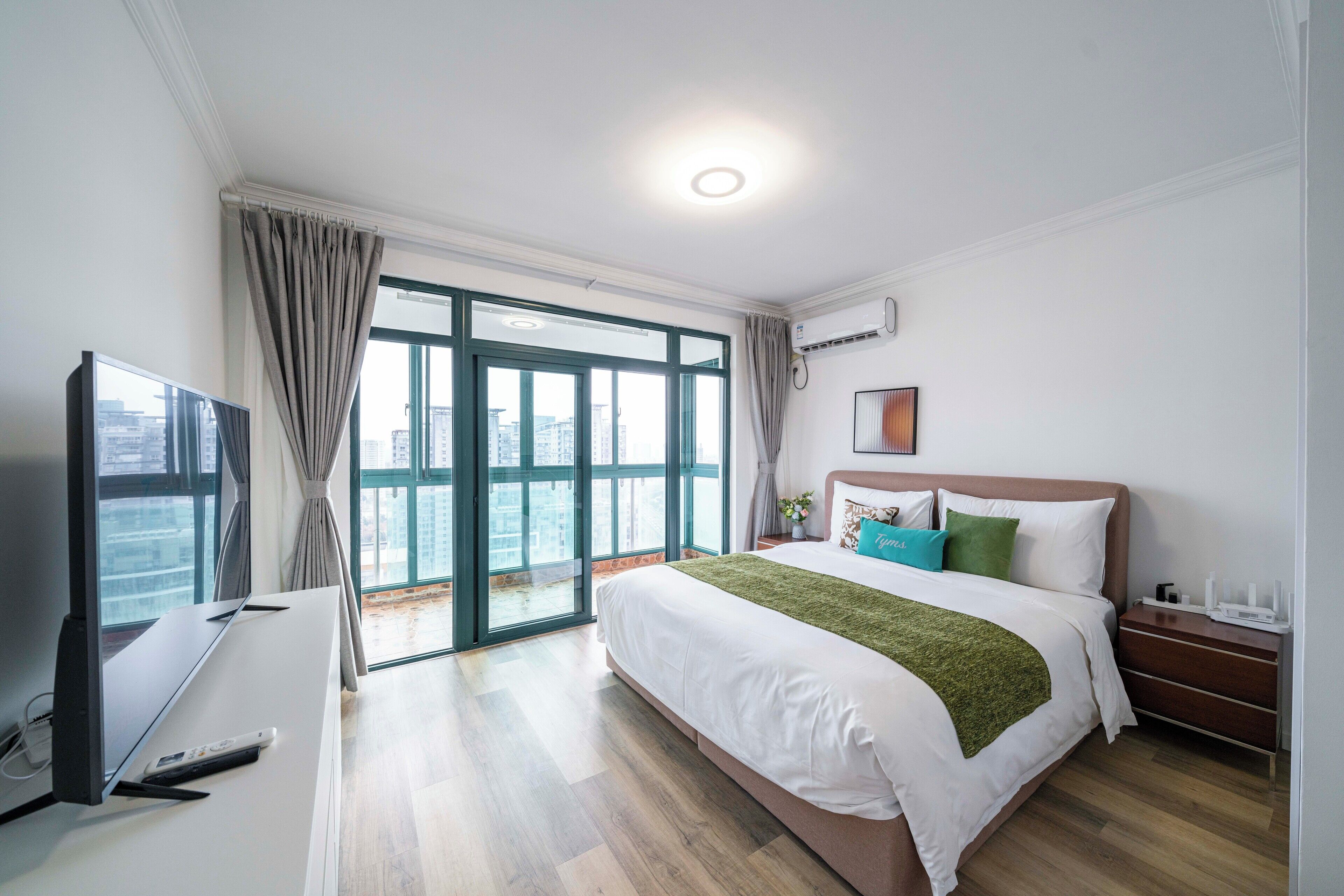 A hotel room with a bed, a TV, a desk, and a view of the city through large windows.