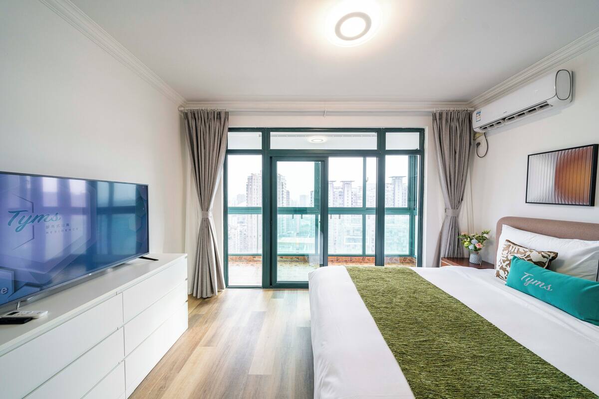 Habitación de hotel con televisor grande, cama con ropa de cama verde y blanca, ventana con cortinas y vistas a la ciudad.