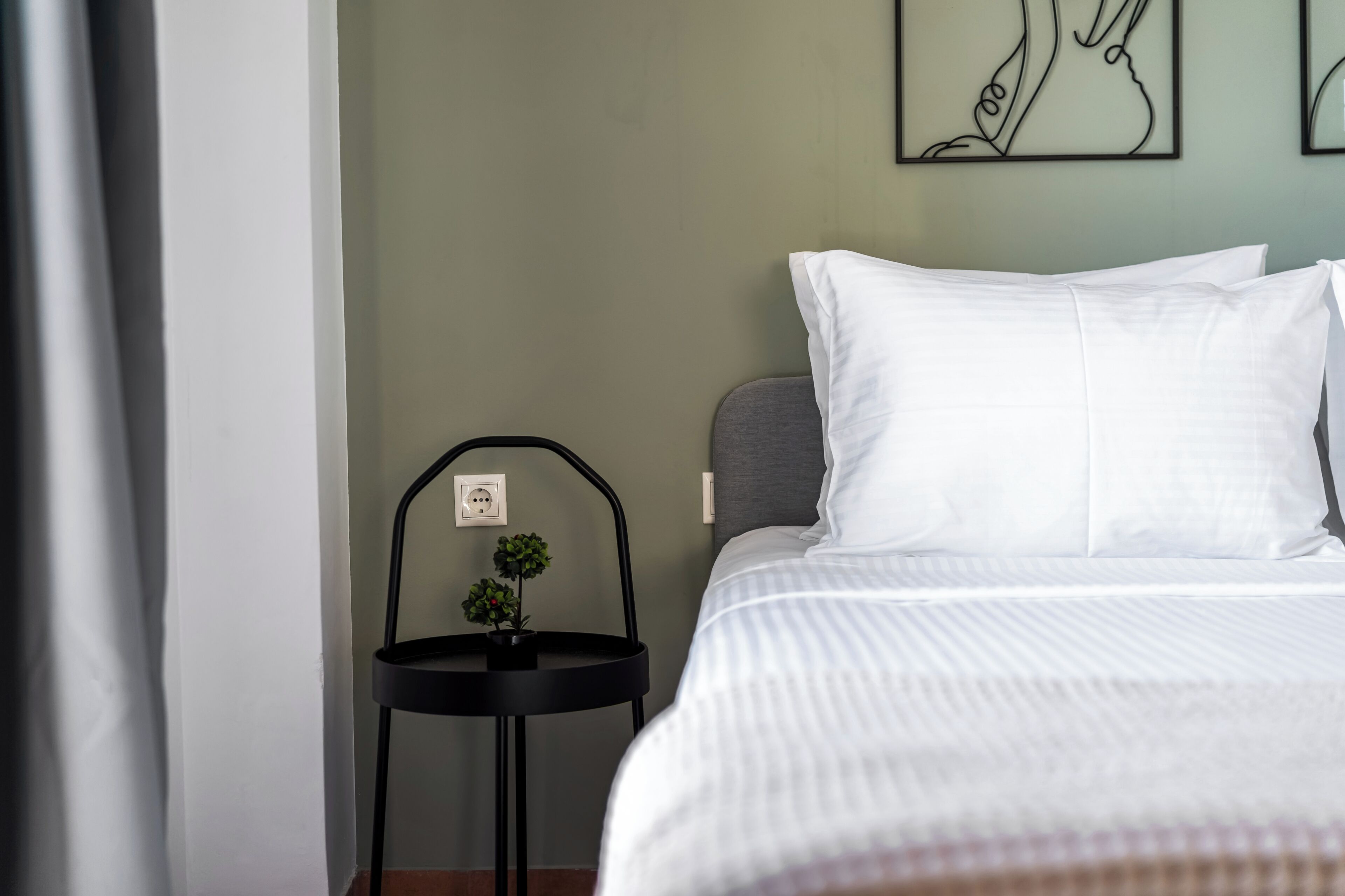 A neatly made bed with white linens, a small black side table with a plant, and a wall with a single artwork.