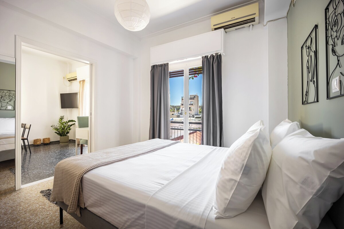 A hotel room with a bed, a TV, a desk, a chair, a potted plant, and a balcony with curtains.