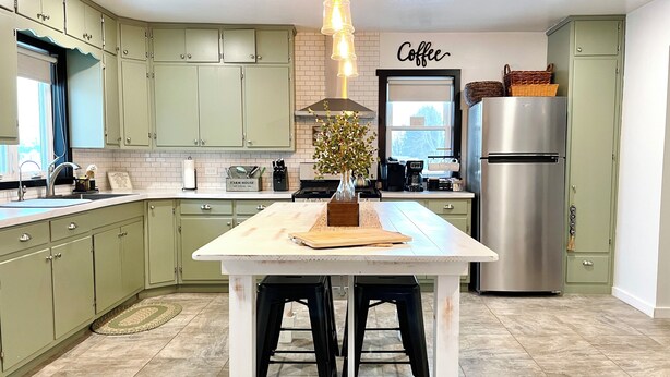 A kitchen with light green cabinets, a white island with stools, a stainless steel refrigerator, and a 'Coffee' sign on the wall.