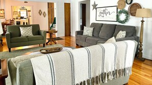 A living room with two sofas, a coffee table, and a lamp. There is a kitchen visible in the background.