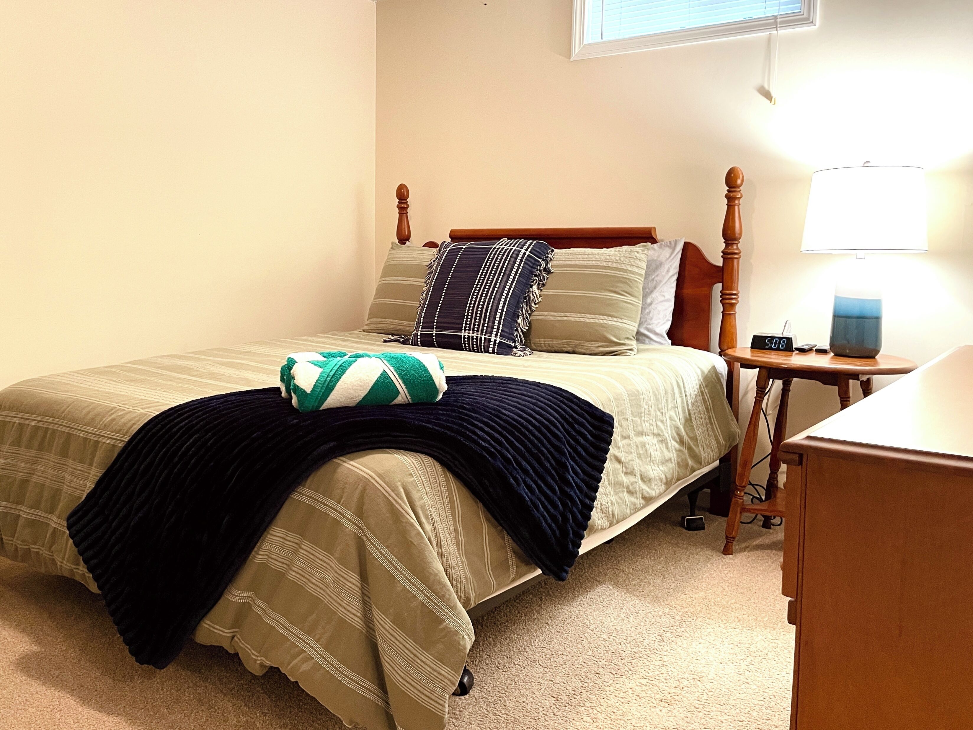 A neatly made bed with a striped blanket and pillows, a bedside table with a lamp and a digital clock, and a window with blinds.