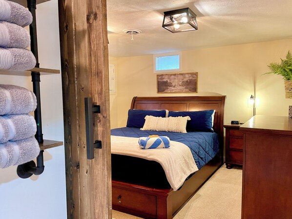 A bedroom with a wooden bed, a nightstand, a wall-mounted shelf with towels, a ceiling light, and a window with blinds.