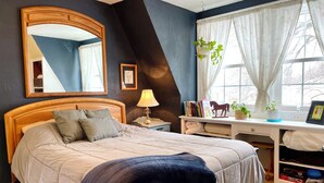 Een slaapkamer met een bed, nachtkastje, lamp, boeken en een raam met gordijnen.