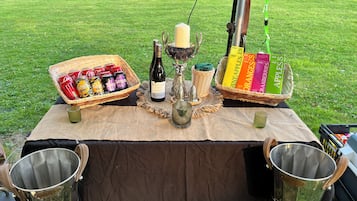 Preparació per a un pícnic amb una taula coberta amb un drap marró, que inclou una ampolla de vi, una espelma i cistells plens d'aperitius i begudes.