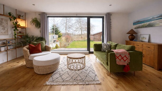 Ein Wohnzimmer mit einem grünen Sofa, einem runden Kaffeetisch aus Weidwerk und einem Korbstuhl. Es gibt eine Holzkommode und ein Bild an der Wand.
