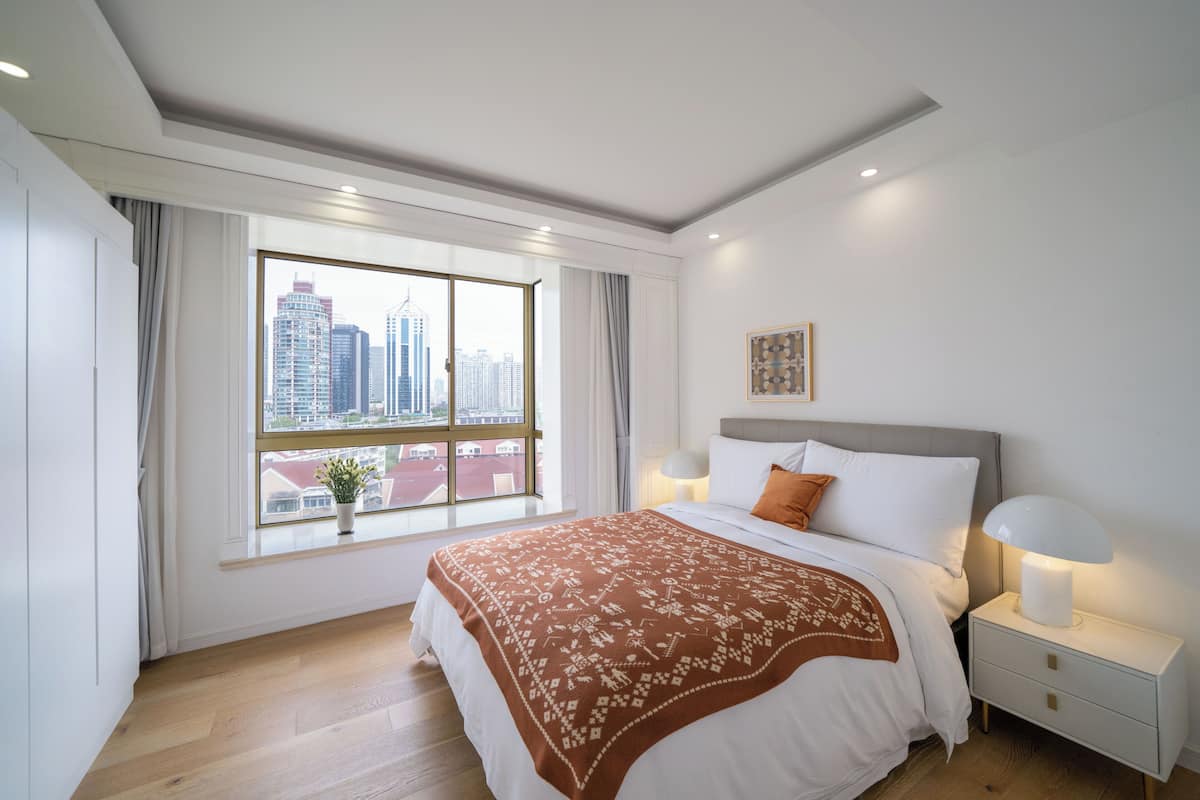 Une chambre avec une grande fenêtre, un lit recouvert d’une couverture à motifs, une lampe de chevet et une table de chevet.