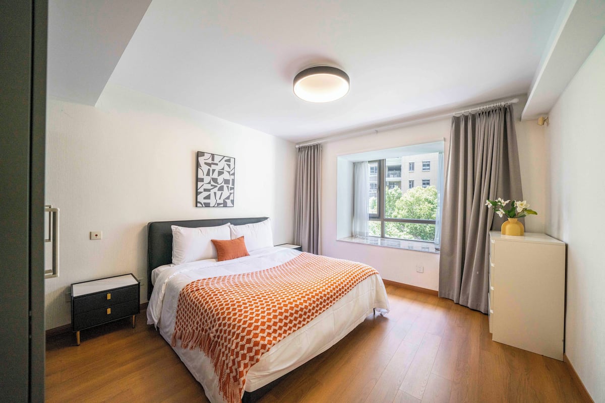 Une chambre à coucher comprenant un lit, une table de chevet, une commode, une fenêtre avec des rideaux et un luminaire au plafond.