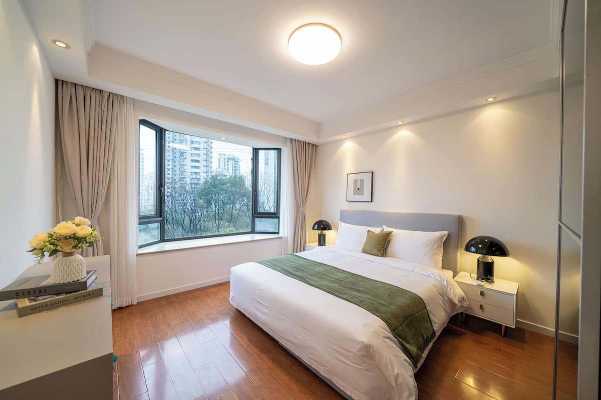 Une chambre d’hôtel moderne avec un grand lit, des lampes de chevet, une table de nuit, une fenêtre avec des rideaux et une vue sur les bâtiments environnants.