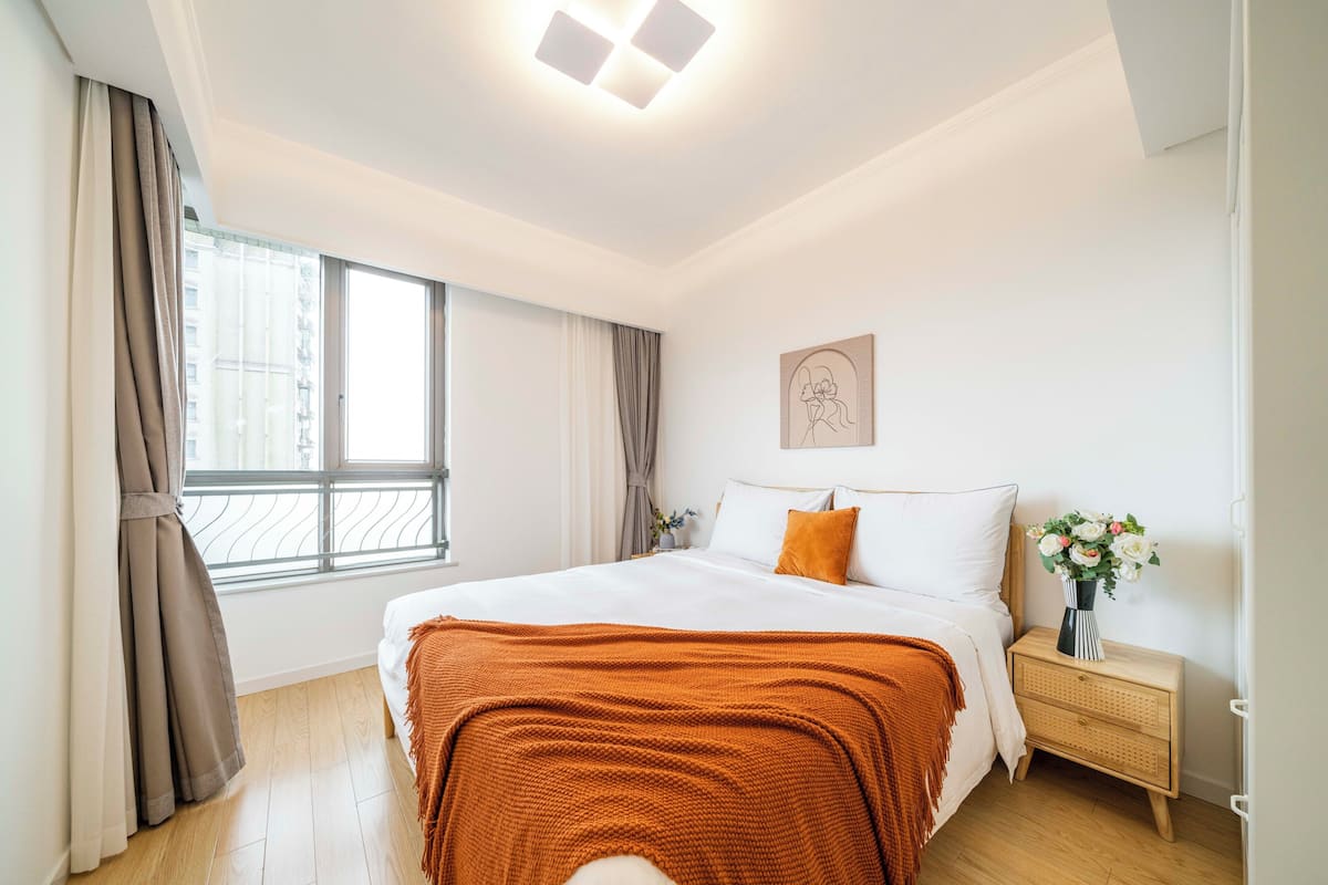 Une chambre à coucher avec un grand lit, une table de chevet ornée d’un vase de fleurs et une fenêtre avec des rideaux.