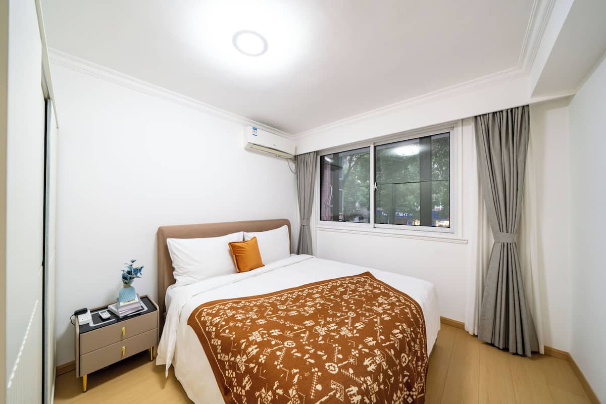 Une chambre à coucher avec un lit, une table de chevet, une commode et une fenêtre avec des rideaux.