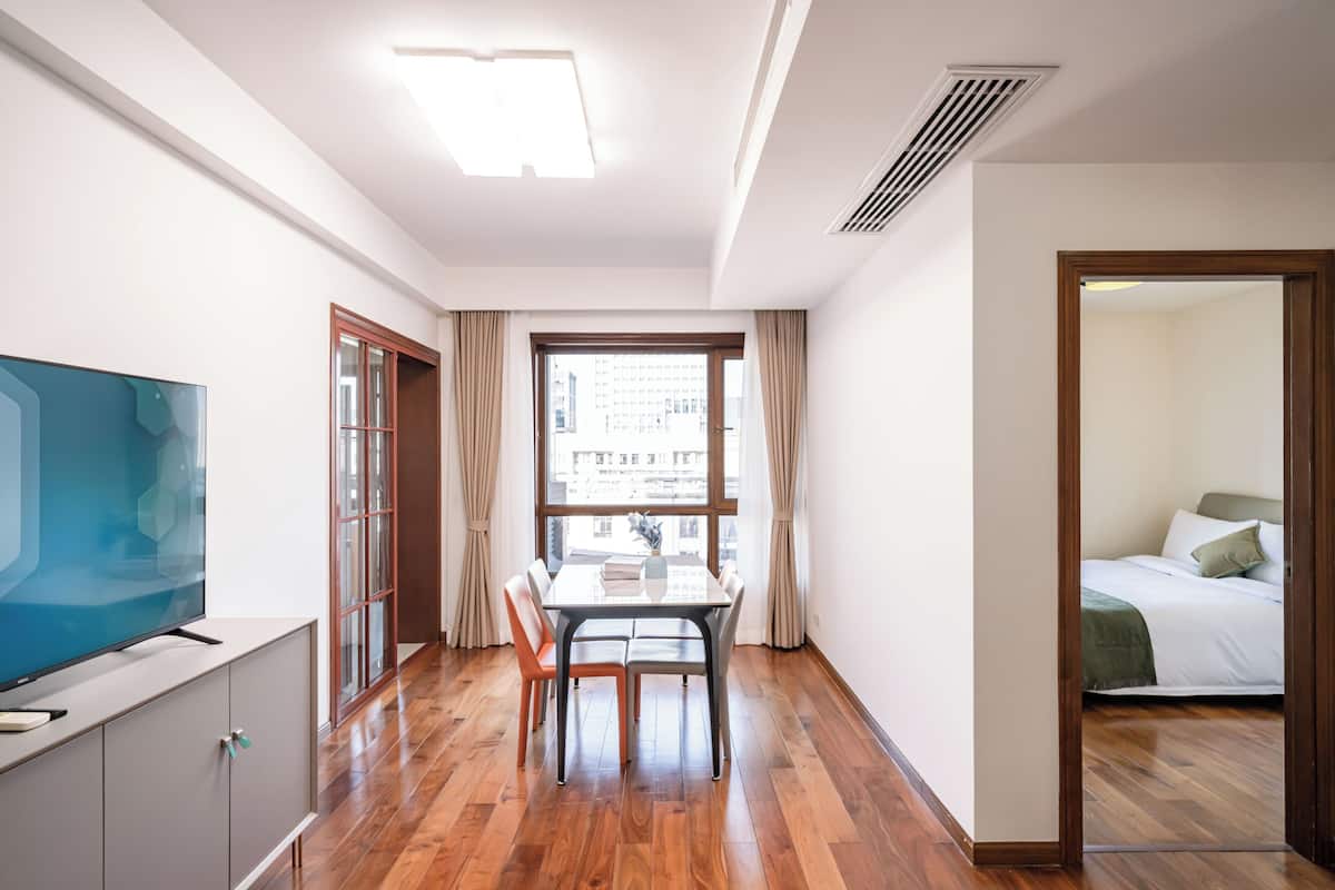 Une chambre d’hôtel moderne avec un plancher en bois, un coin repas comprenant une table et des chaises, un téléviseur posé sur un meuble et une grande fenêtre avec des rideaux.