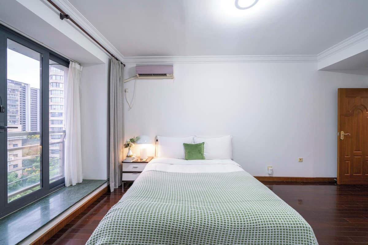 Une chambre avec une grande fenêtre, un lit avec un oreiller vert, une table de chevet avec une lampe et une porte en bois.