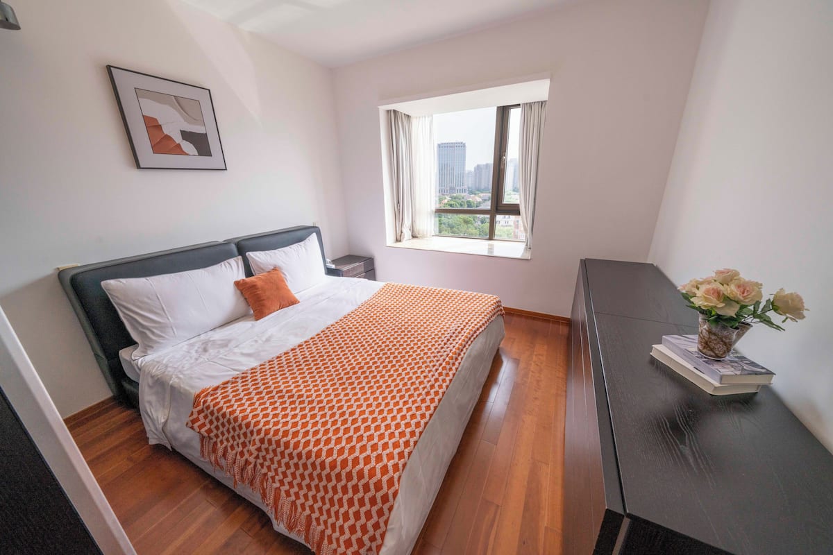 Une chambre d’hôtel avec un lit, une table de chevet, une fenêtre avec des rideaux, un tableau encadré au mur et un vase avec des fleurs sur une table.