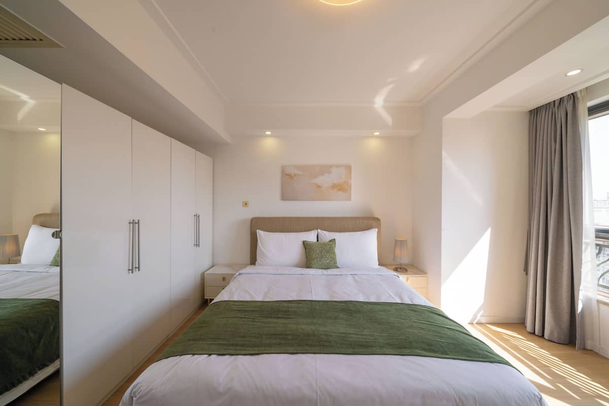 Une chambre d’hôtel avec un lit, une table de chevet, un miroir, une fenêtre avec des rideaux et un tableau au mur.