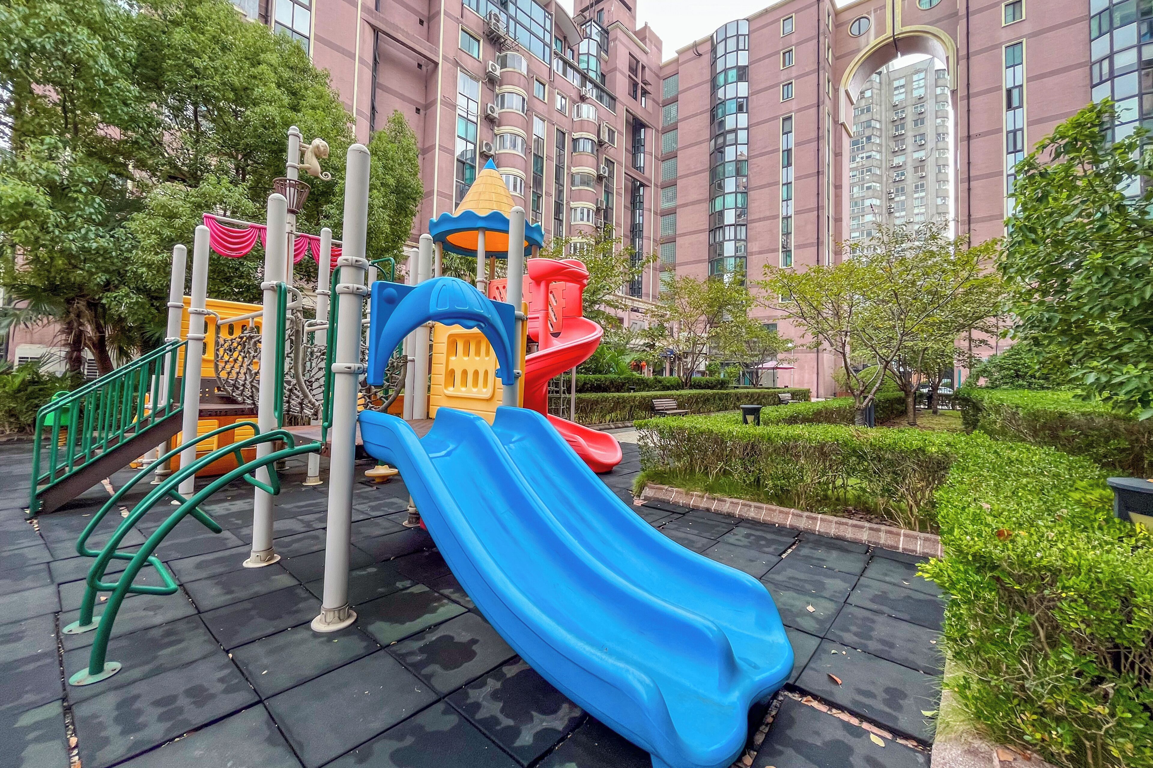 A playground with slides and climbing structures, surrounded by greenery and a multi-story building in the background.