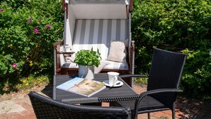 Um pátio com jardim, uma poltrona listrada, uma pequena mesa com um livro e uma chávena, e um vaso com planta.
