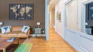 A living room with a brown leather sofa, a wooden coffee table, and a green patterned rug. There are two small side tables with lamps. A world map artwork is on the wall. - Welcome to Colosseum Haven Steps from Roman Forum (Roma)