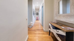 A hallway with a wooden floor, a white wall with a decorative mirror, and a dog sitting on the floor. - Welcome to Colosseum Haven Steps from Roman Forum (Roma)